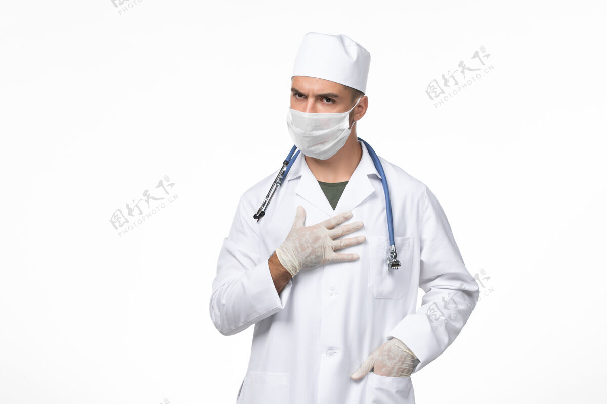 佩戴正面图：男医生穿着医疗服 戴着防冠状病毒口罩 浅白色墙壁上有蓝色听诊器 冠状病毒-疾病大流行病毒正面查看西装