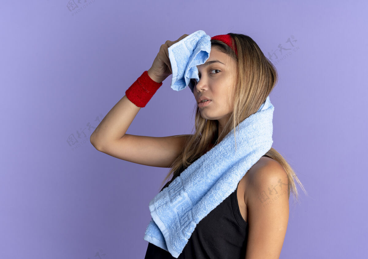 年轻身穿黑色运动服 头戴红色头带 头戴耳机的年轻健身女孩站在蓝色的墙上用毛巾擦额头立场健身耳机