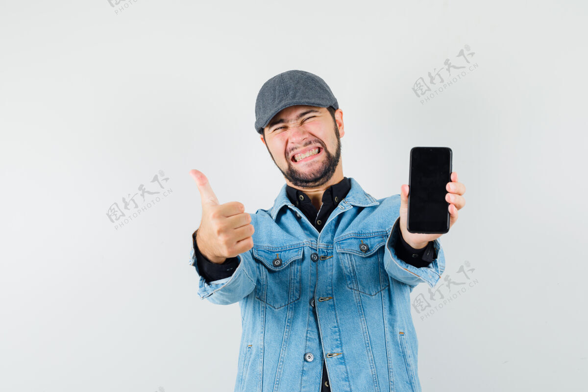 夹克复古风格的男人展示拇指向上 同时显示手机在夹克 帽子 衬衫和看起来很高兴 前视图老年轻成人