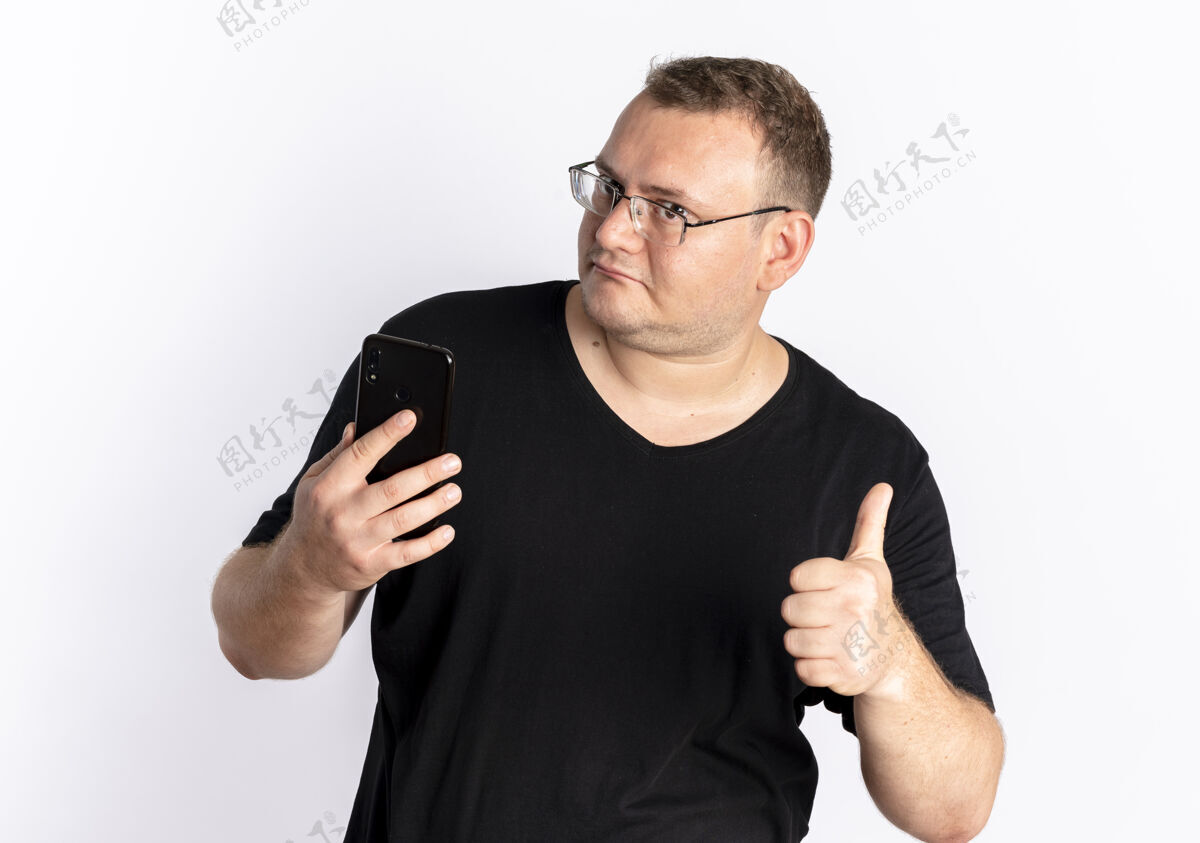站一个戴眼镜 身穿黑色t恤的超重男子站在白色的墙上 看着他的智能手机屏幕 竖起大拇指拇指男人屏幕