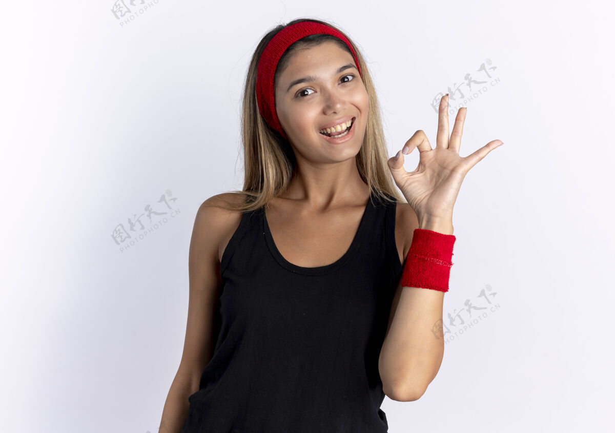 健康身穿黑色运动服 头戴红色头巾的年轻健身女孩微笑着站在白色墙壁上展示ok标志女孩运动装微笑