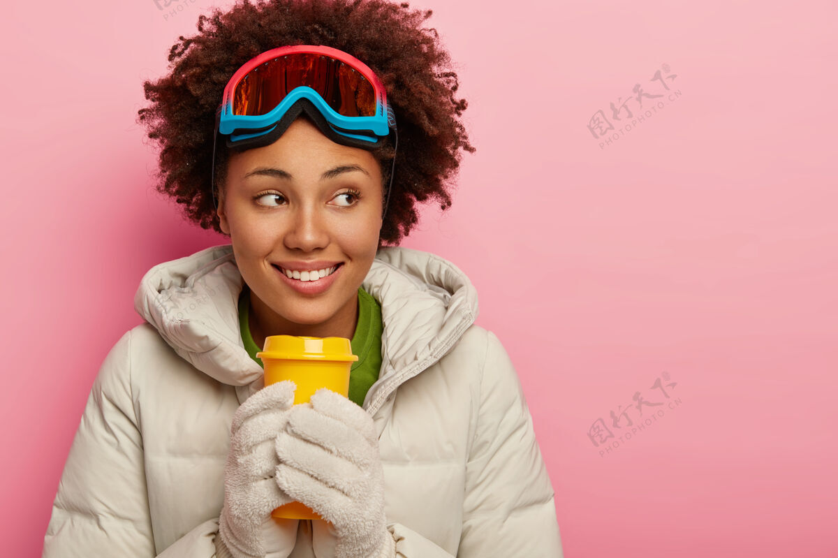 喝酒微笑的卷发女人拿着外卖咖啡 用热饮料取暖 穿着白色冬衣和滑雪眼镜护目镜室内手套