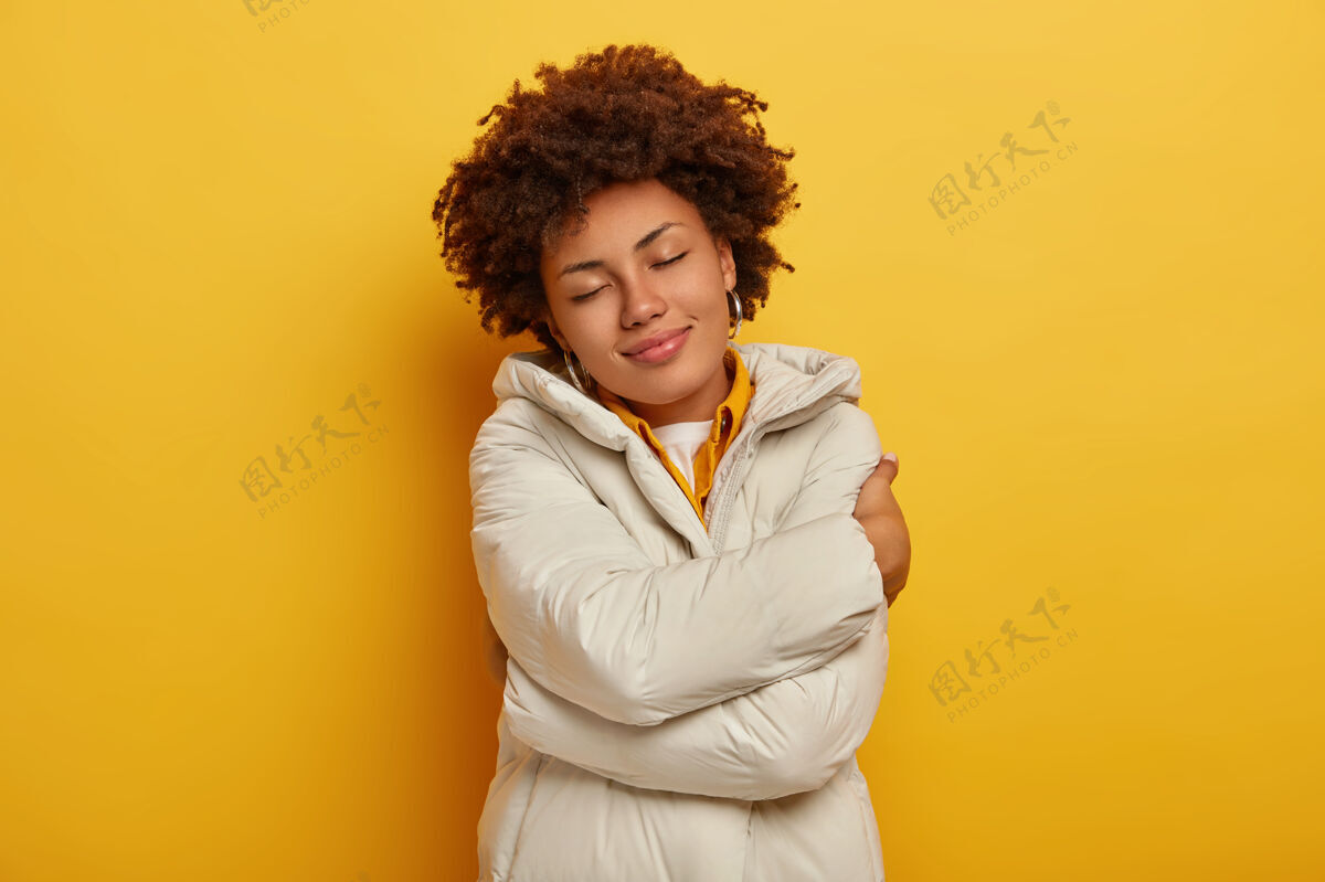 休闲美丽快乐的女人穿着新的冬季夹克享受舒适 拥抱自己 闭上眼睛 感到温暖和满足 头发卷曲 黄色背景下孤立人 衣服的概念黄色年轻白色