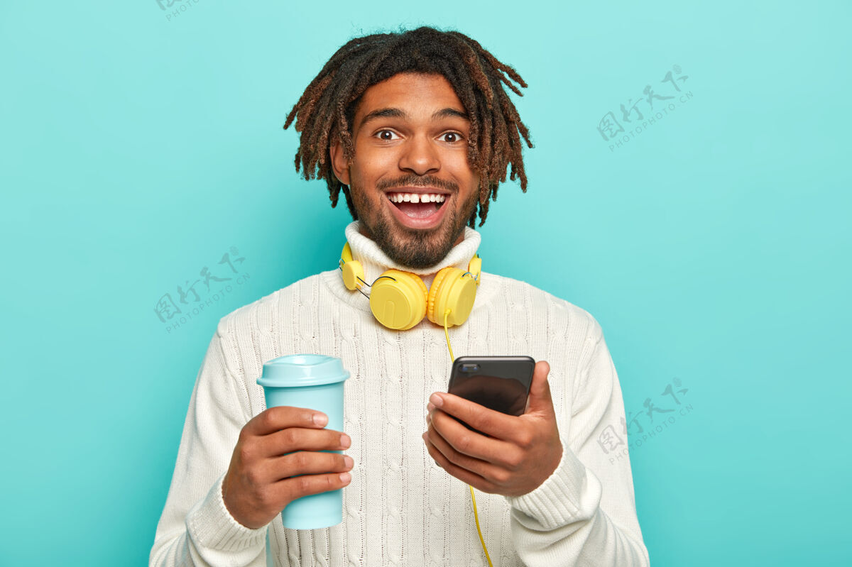 耳机帅气开朗的男人的照片 留着可怕的长发 拿着现代手机和外卖咖啡 带着耳机微笑杯子一个