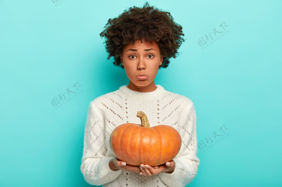 庆祝摄影棚拍摄了一个非洲发型 抱着南瓜 穿着白色针织毛衣 愁眉苦脸地看着镜头的困惑的年轻女子毛衣困惑蔬菜