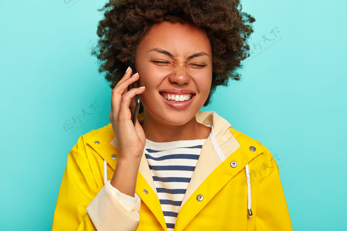 外套剪短的形象 迷人的黑皮肤女人与非洲头发 笑有趣的朋友的故事 拿着手机 穿着黄色雨衣雨衣讨论持有