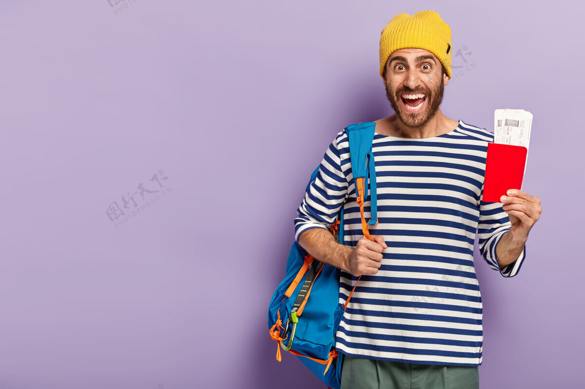 举行快乐的未刮胡子男背包客的摄影棚镜头拿着护照和证件 肩上扛着背包 高兴地笑着 穿着休闲装 隔着紫色的墙壁 准备旅行水平留茬黄色
