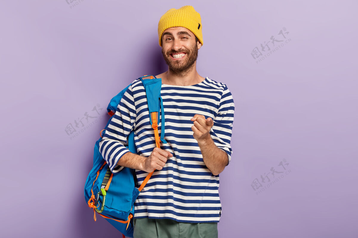 一个快乐微笑的男人旅行者的照片用食指指着你 背着背包 戴着黄色帽子和毛衣 表达选择 接你 孤立在紫色的墙上年轻徒步旅行者白种人