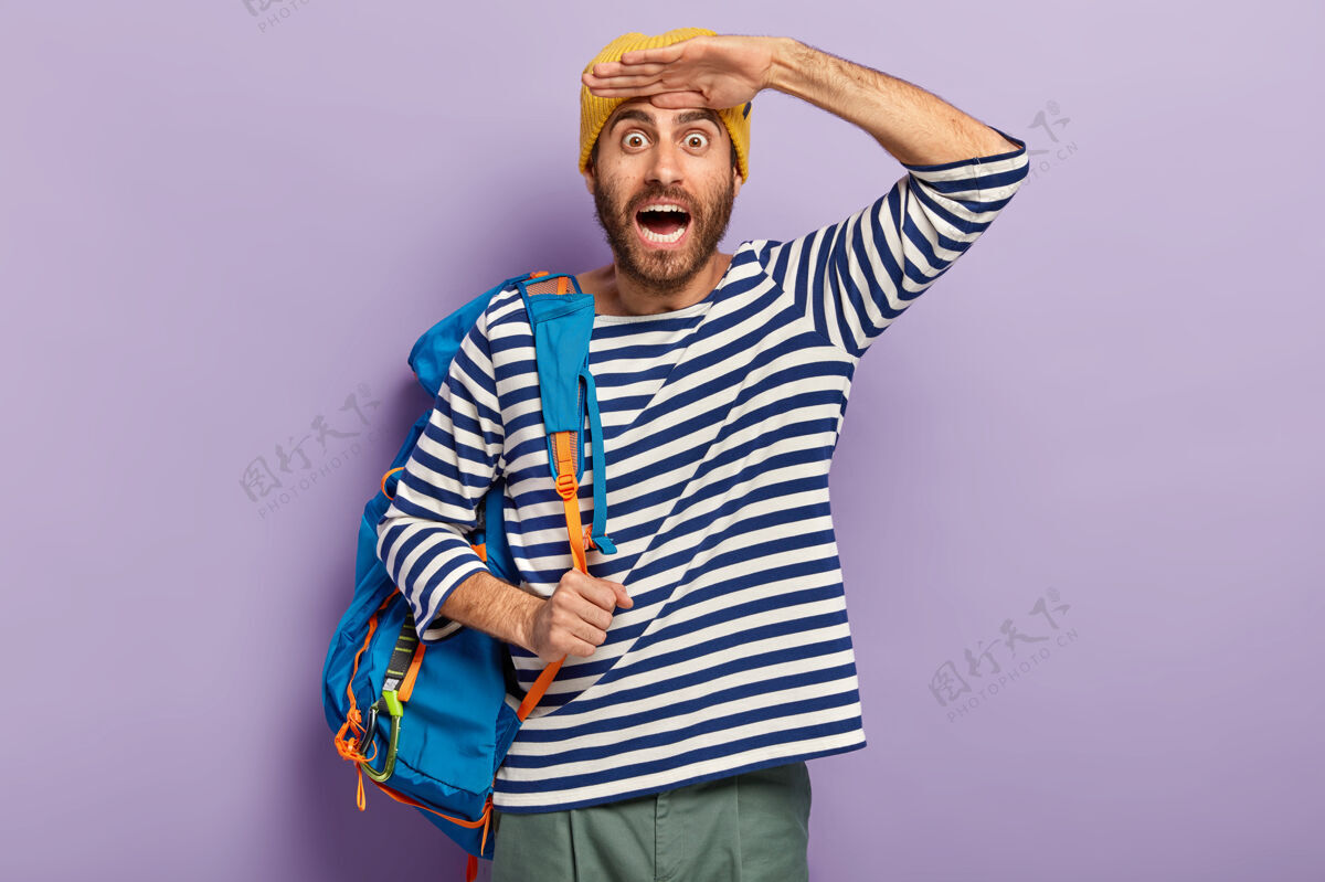 距离情绪激动惊喜开朗的年轻男性游客手心紧靠额头 穿着条纹毛衣 背着蓝色背包 里面装着私人物品蓝色紫色徒步旅行
