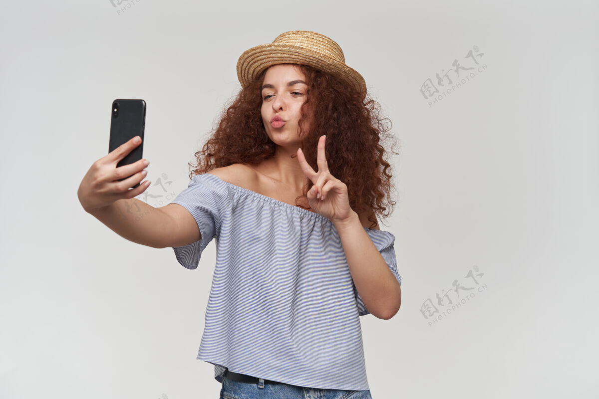 女士十几岁的女孩 快乐的女人 卷曲的姜黄色头发穿着条纹露肩衬衫和帽子在智能手机上自拍 展示和平标志和亲吻在白色的墙壁上孤立地站着看姜服装