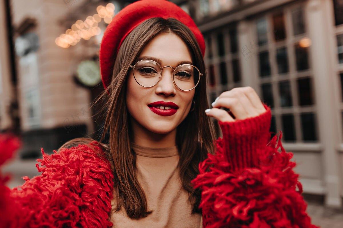 羊毛红唇美女在街上自拍戴着时髦帽子 红色夹克和米色上衣 戴着眼镜的黑发女郎的照片美丽年轻阳光