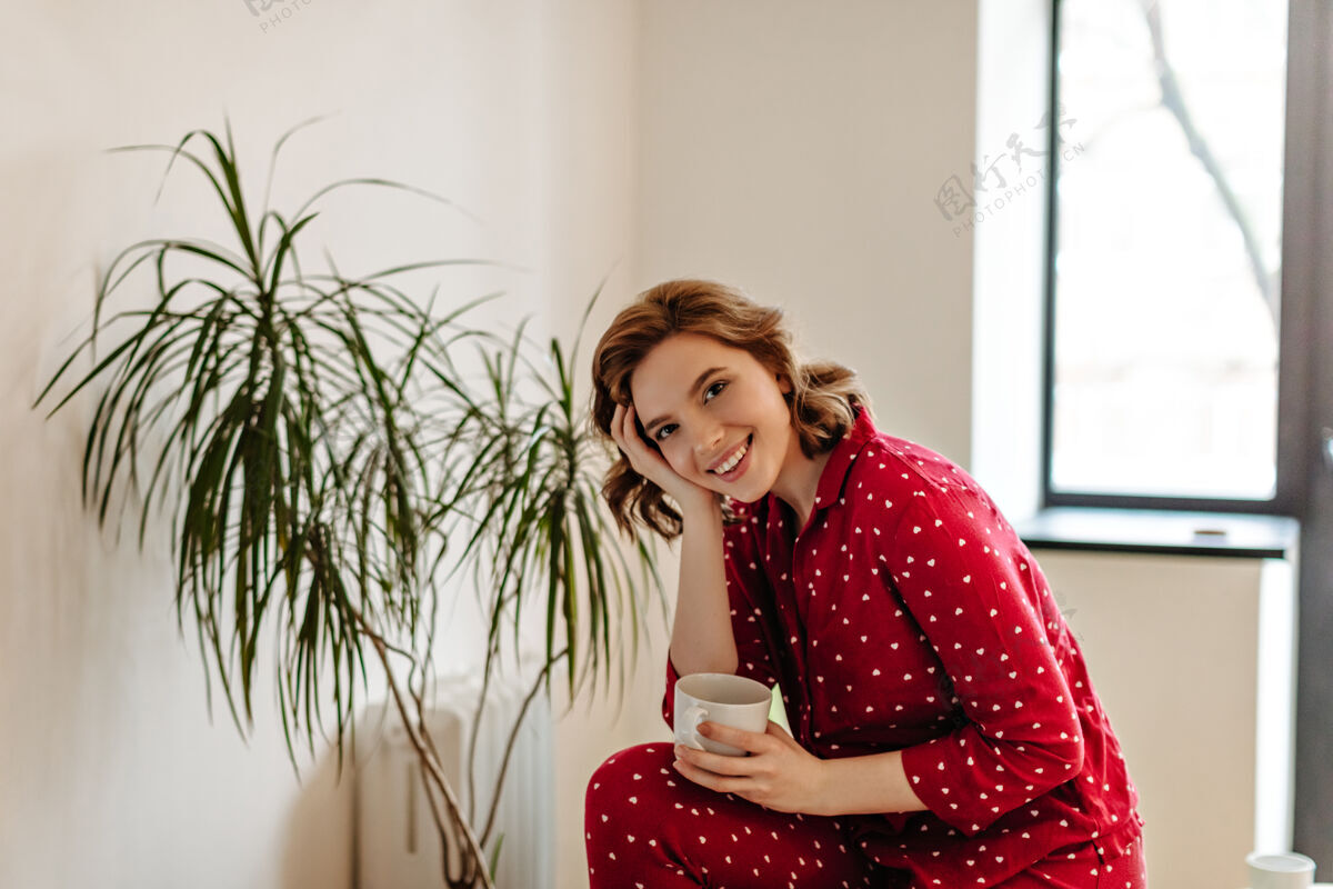 放松穿着睡衣喝着茶的梦幻女人的室内镜头拿着一杯咖啡微笑的白人年轻女子房子无忧无虑饮料