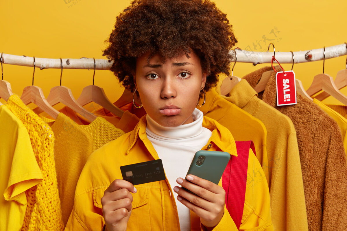 沮丧失意女买家逛服装店 用手机和信用卡进行网上支付 站在黄色背景下的照片设备不满意支付