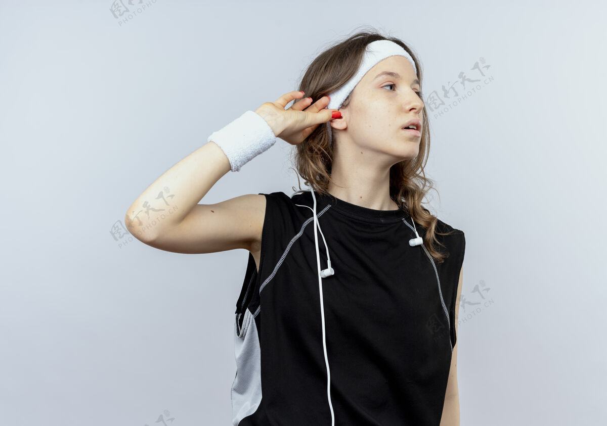 尝试身穿黑色运动服的年轻健身女孩戴着头巾 站在白墙上 一边看着一边抚摸着耳朵 试图倾听一边年轻健身