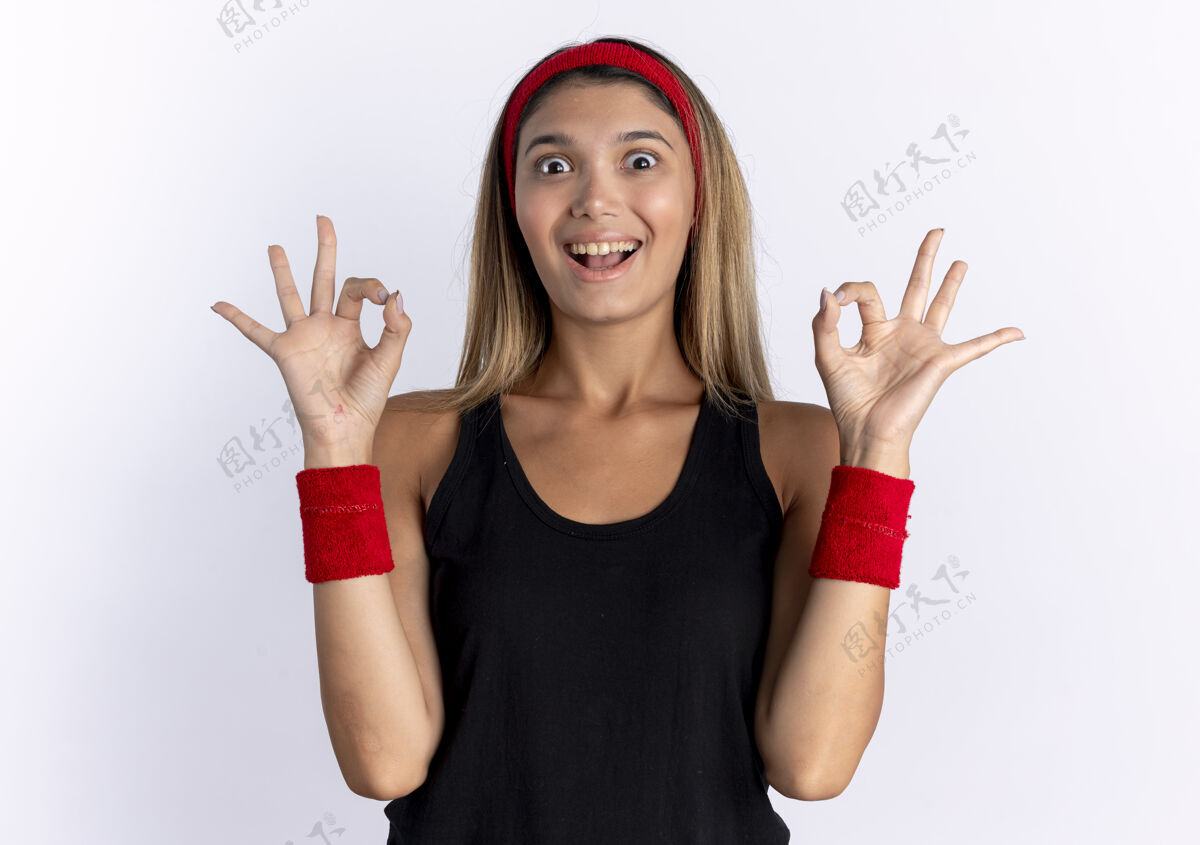 好身穿黑色运动服 头戴红色头巾的年轻健身女孩微笑着站在白色墙壁上展示ok标志站秀运动装
