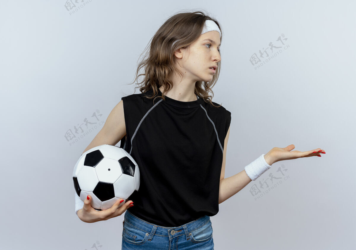 女孩身穿黑色运动服的年轻健身女孩 头上戴着足球带 手臂伸出 站在白色的墙壁上 一边看着一边问道头带运动装年轻