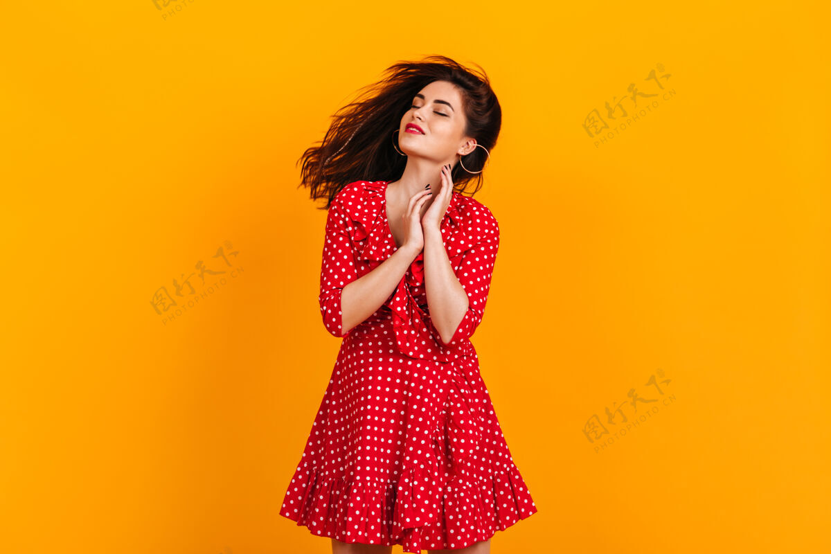 高跟鞋身着红色圆点装的年轻女子微笑着闭着眼睛在黄色的墙上摆姿势女性魅力站立