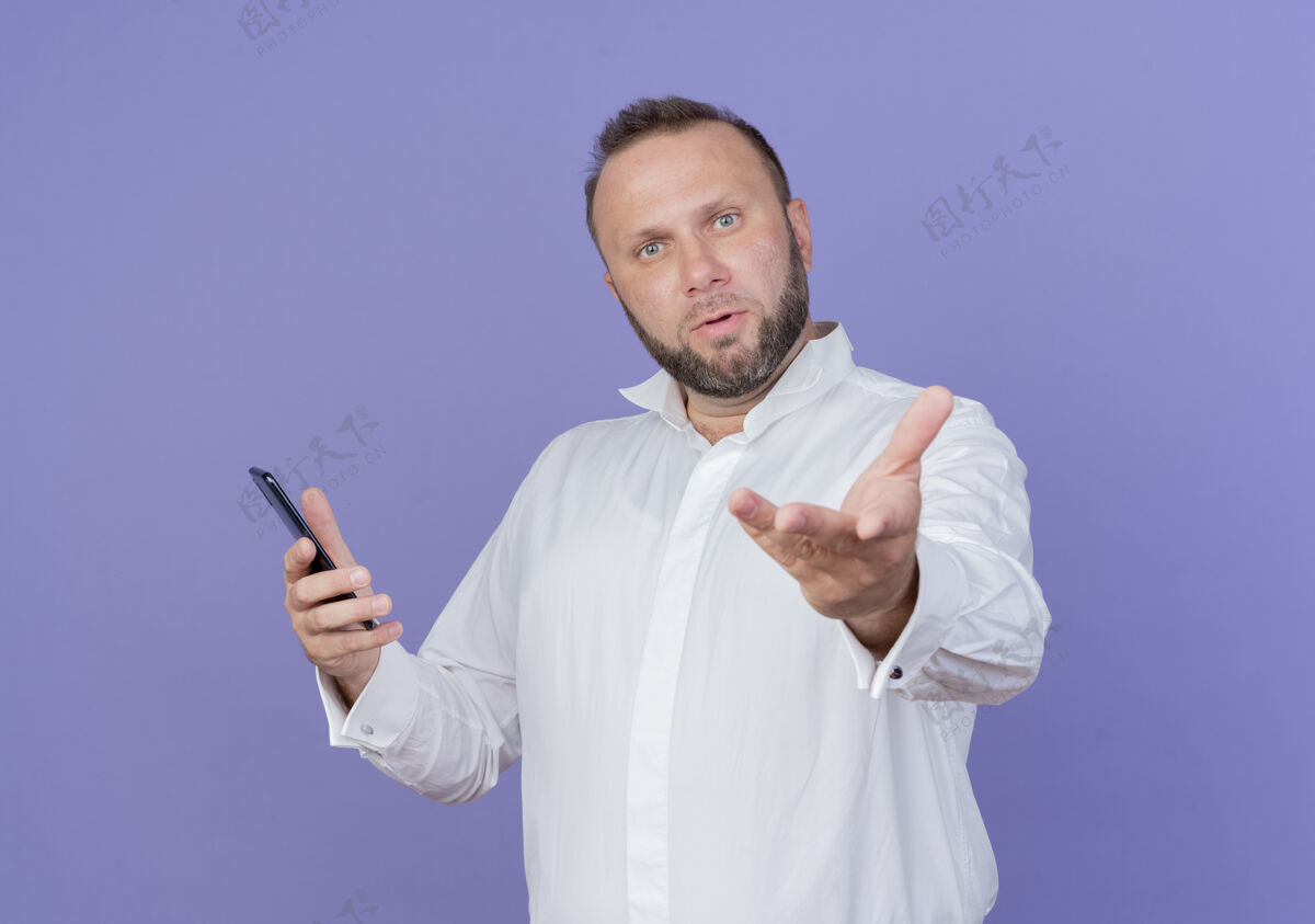 问一个留着胡子的男人穿着白衬衫 拿着智能手机 伸出胳膊 站在蓝色的墙壁上 问自己困惑胡子穿抱着