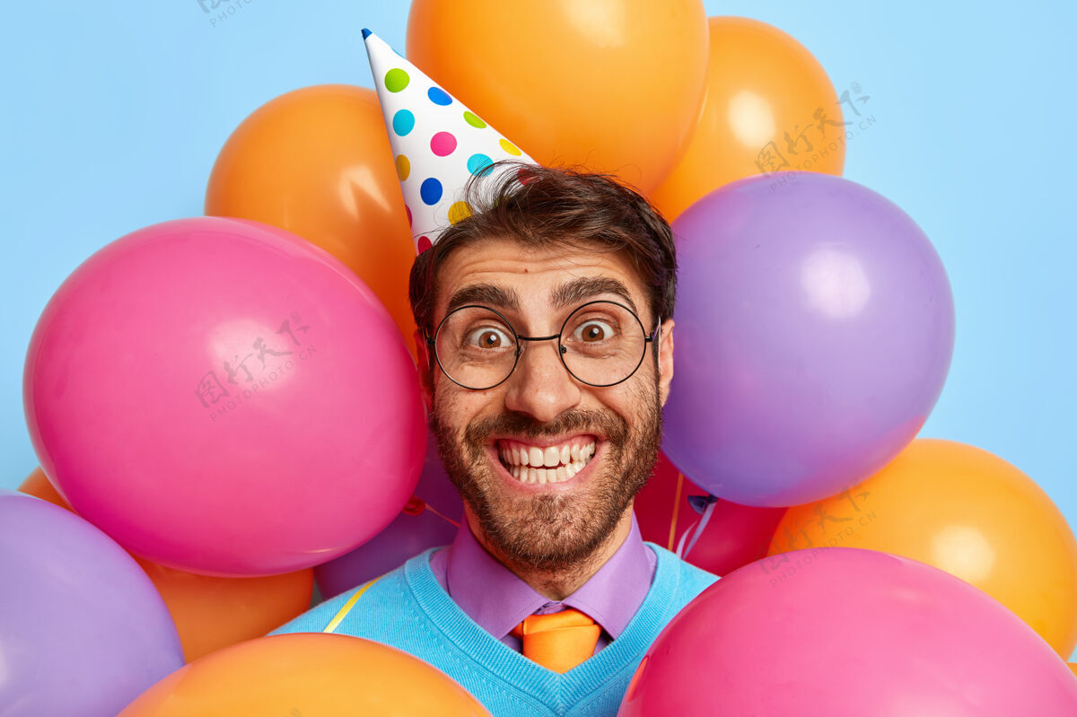 完美帅哥快乐的头像被派对气球包围着摆姿势帽子眼镜男人