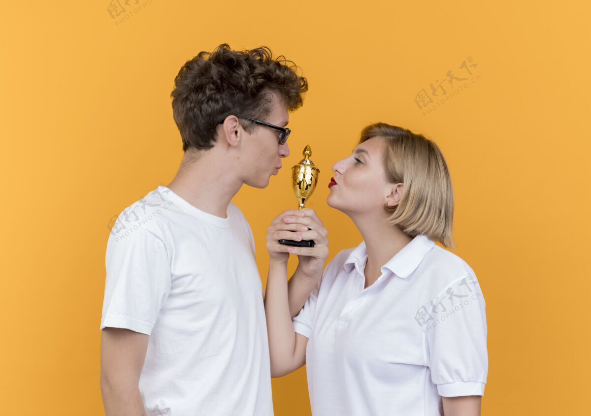 女人一对年轻的运动情侣手拿奖杯站在一起看着对方微笑着亲吻他们站在橙色墙上的奖杯看奖杯运动