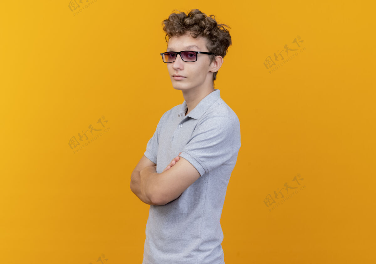 胸部戴着黑眼镜的年轻人 穿着灰色马球衫 严肃的脸 双手交叉放在胸前 站在橙色的墙上穿男人眼镜