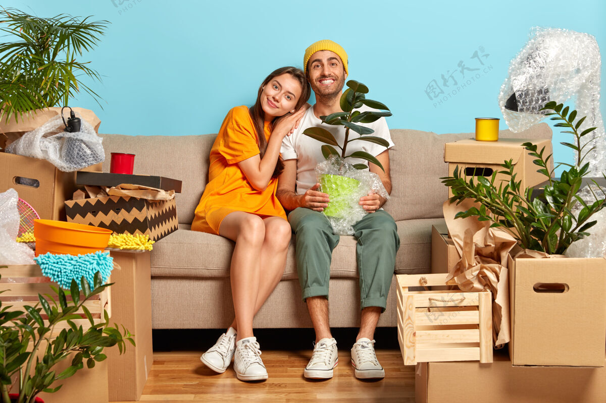 丈夫微笑的年轻夫妇坐在沙发上 四周都是盒子室内植物包装新婚夫妇