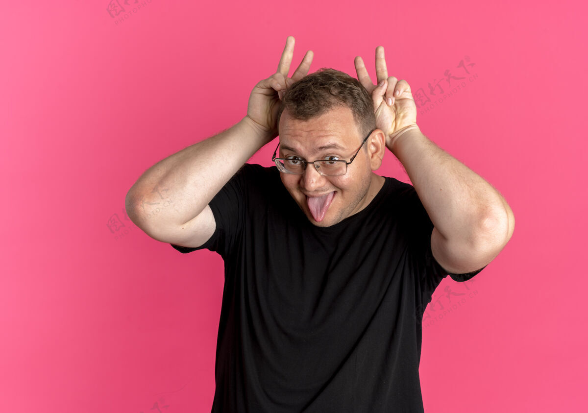 立场一个戴眼镜的胖男人 穿着黑色t恤 一边玩着模仿兔子耳朵伸出舌头站在粉红色的墙上模仿兔子穿