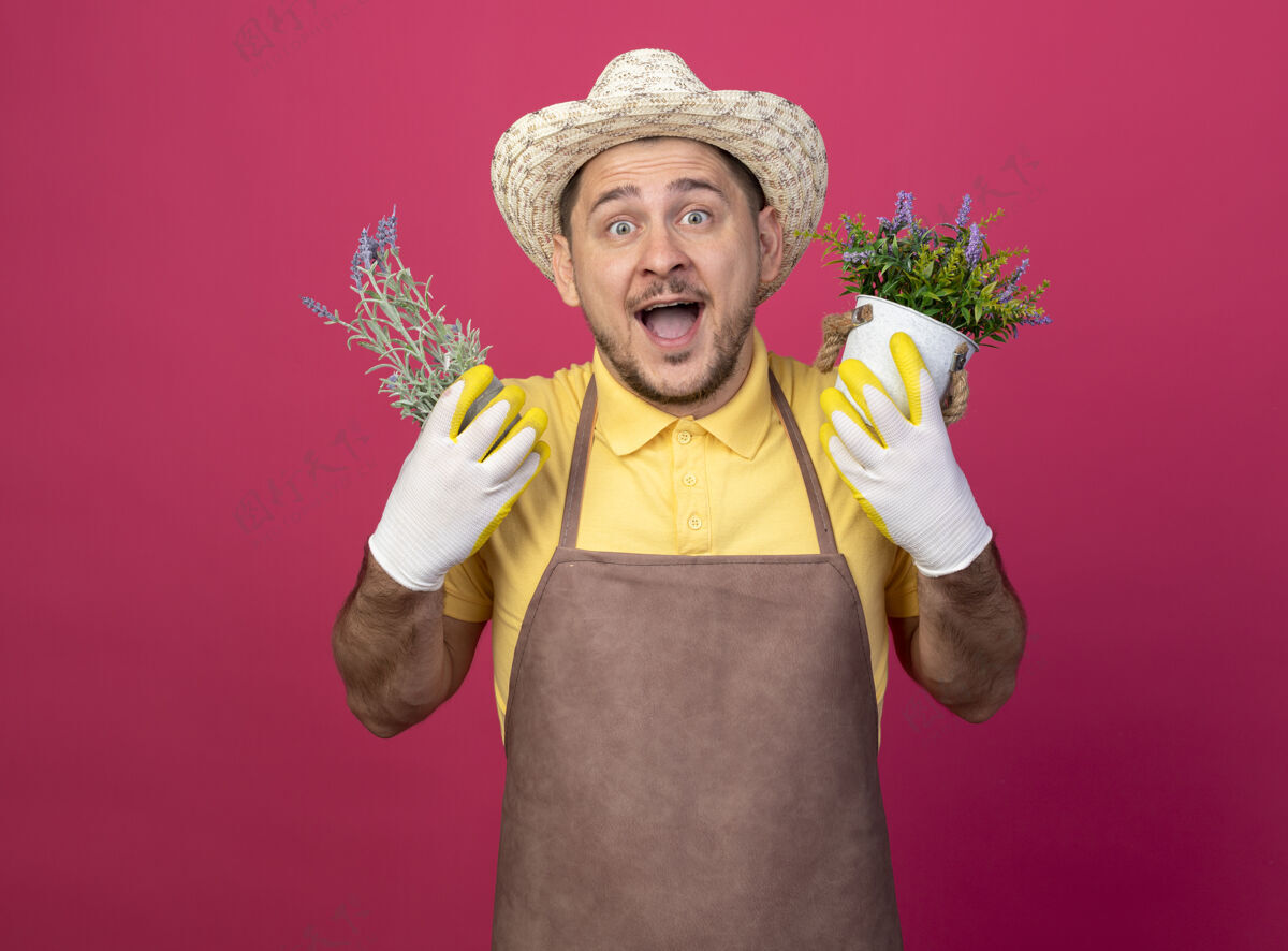 站着年轻的园丁 穿着连体衣 戴着帽子 戴着工作手套 手里拿着盆栽植物 快乐快乐穿手套大麻