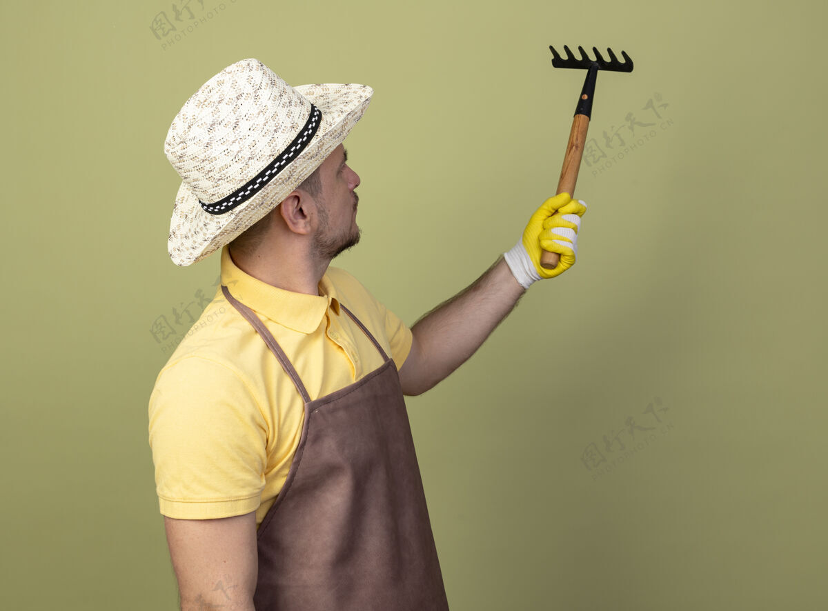 迷你年轻的园丁穿着连体衣戴着帽子戴着工作手套拿着小耙子严肃地看着它耙子男人穿