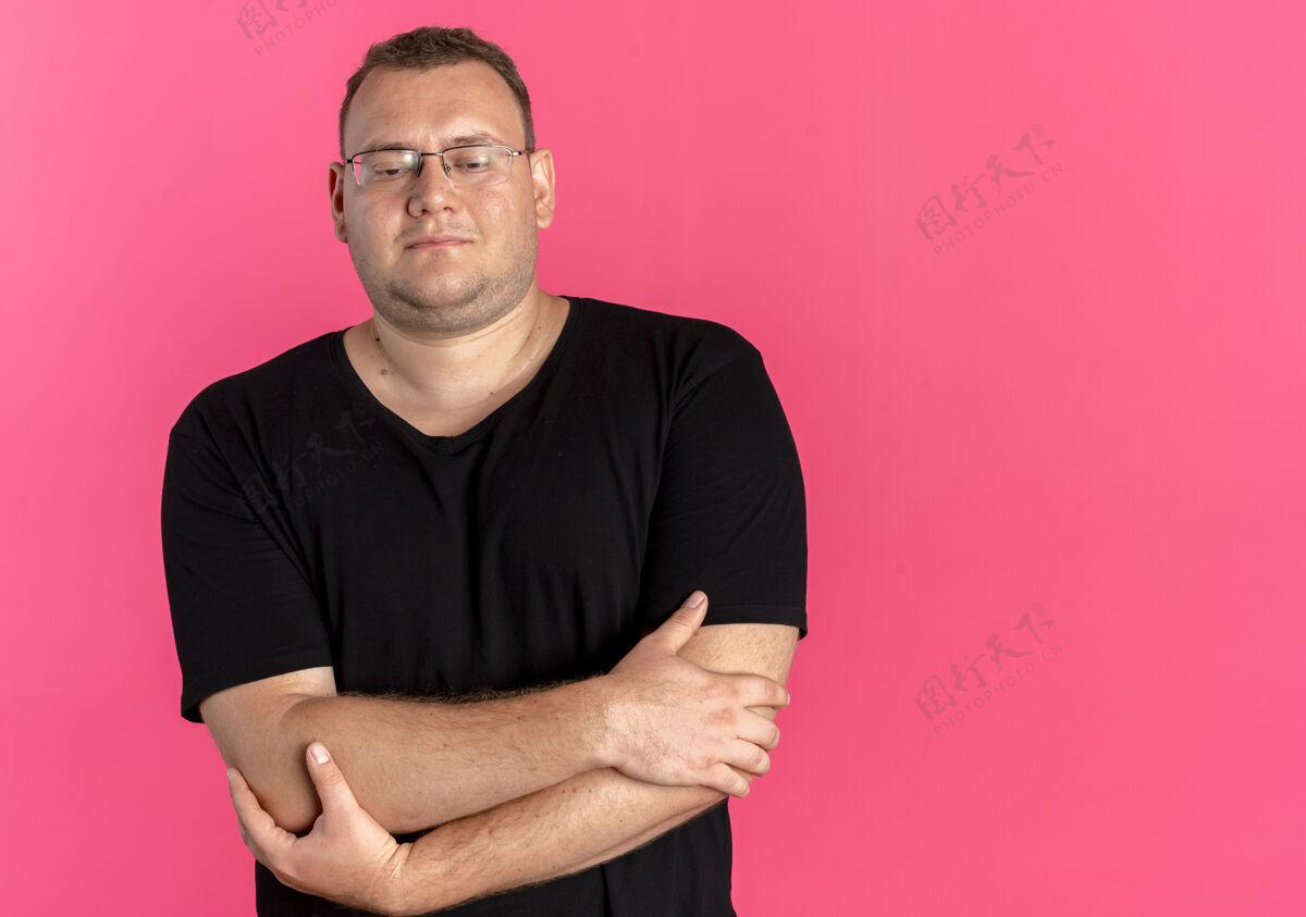 穿一个戴眼镜的超重男人 穿着黑色t恤 脸上带着微笑 双臂交叉在胸前 站在粉红色的墙上脸眼镜男人