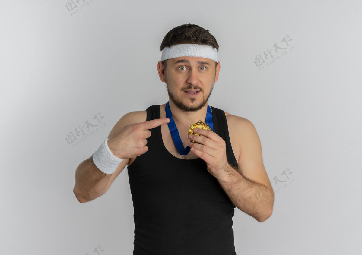 白色戴着头带 脖子上戴着金牌的年轻健身男子站在白色背景下 用手指着金牌 看上去很自信奖牌信心周围