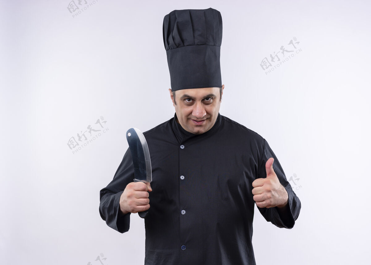 站着男厨师身穿黑色制服 头戴厨师帽 手持锋利的菜刀 微笑着竖起大拇指站在白色背景上显示穿烹饪