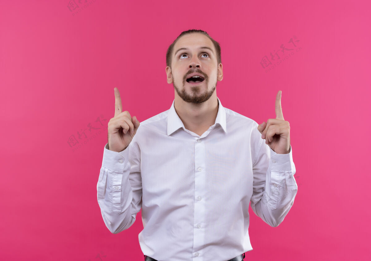 粉色穿着白衬衫的帅哥商人抬起头 用食指指着粉色背景站着 开心而积极衬衫目录正面