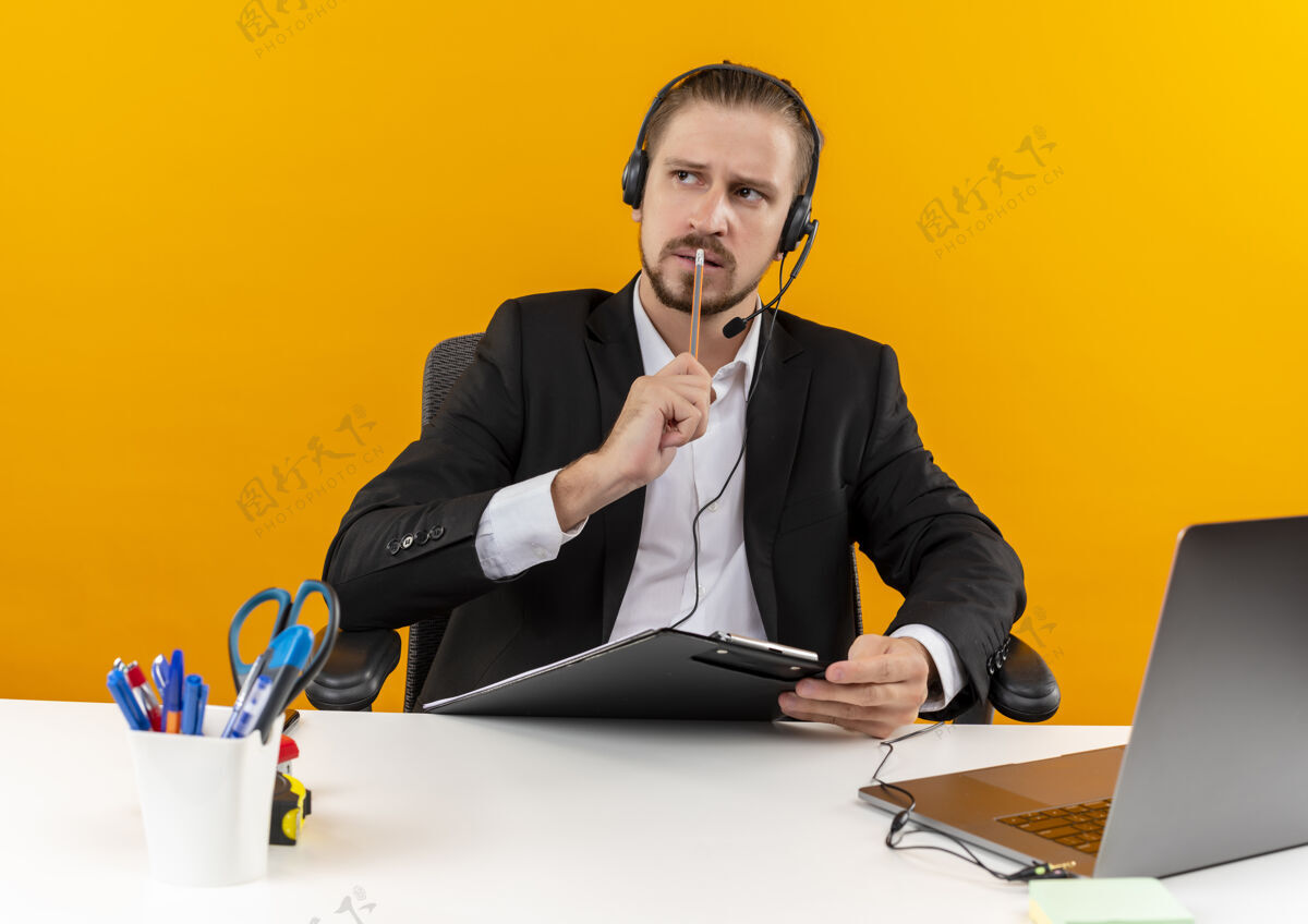 桌子穿着西装戴着耳机拿着麦克风拿着剪贴板困惑地坐在橙色背景下的办公桌旁看着旁边的帅哥麦克风男人坐着