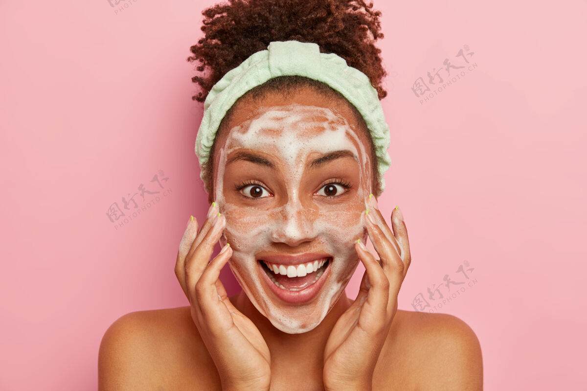 人类笑容可掬的黑皮肤年轻女子把肥皂放在脸上清洗 用冷水洗脸 要有健康新鲜的皮肤 戴上头巾 神采奕奕 赤身裸体站在室内卷发泡沫女性
