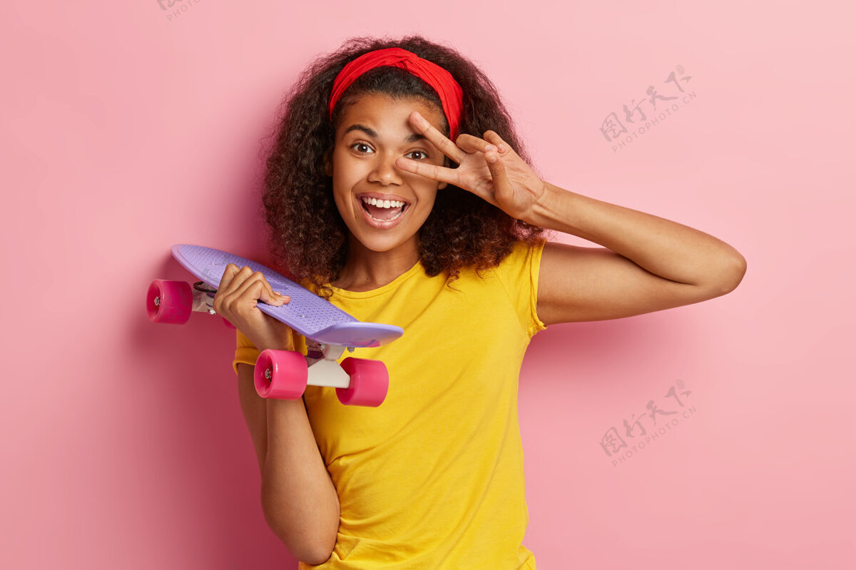 无忧无虑帅气清爽的非裔美国女性头像做出和平姿态 积极微笑 戴着红色头带和黄色t恤 把滑板隔着粉色的墙壁休闲理念人风格欢乐