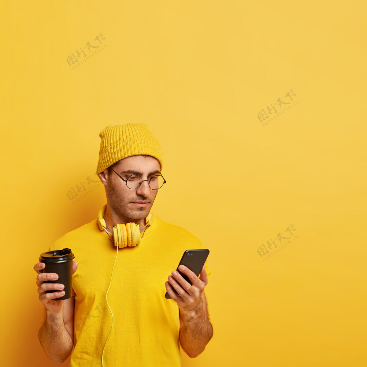 杯子严肃专注的家伙拿着手机 上网看新闻 戴着黄色帽子和t恤 享受着芳香的饮料 连接着免费的wifi手机姿势耳机