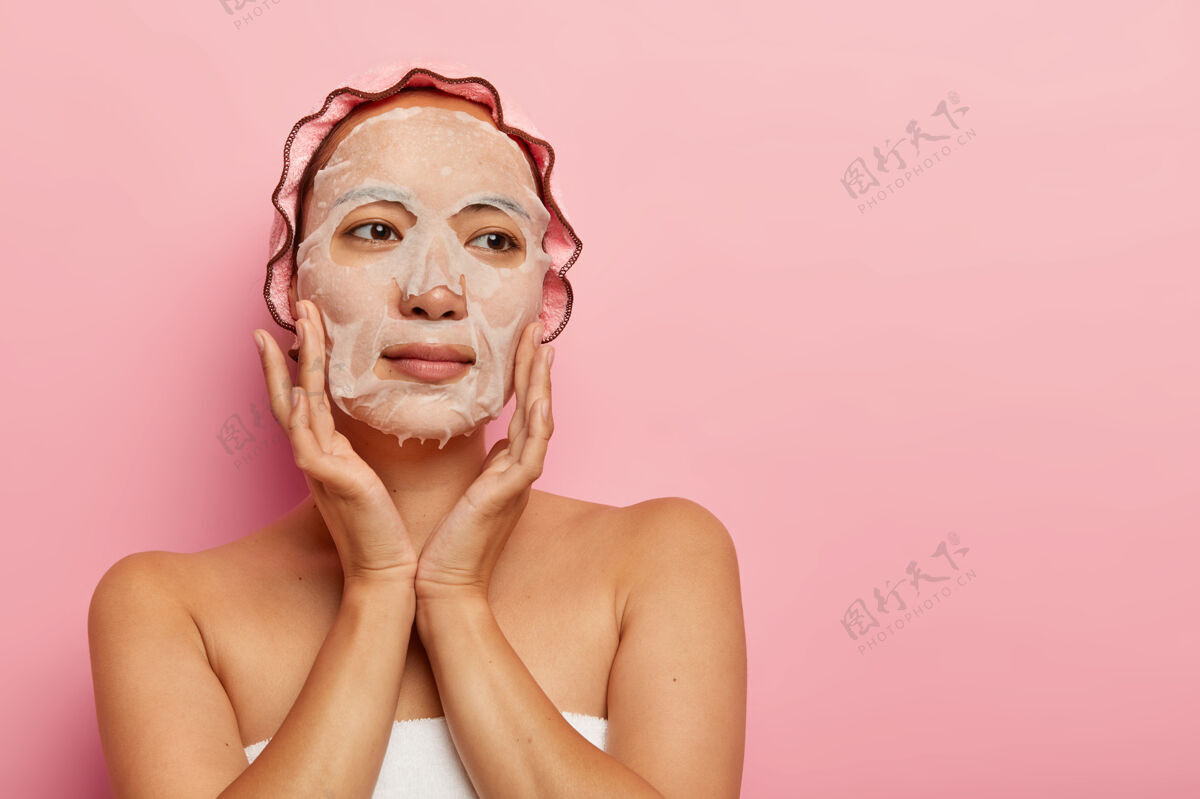 化妆品露肩严肃的女人在脸上敷上保湿纸面膜 轻轻抚摸脸颊 拥有柔软健康的皮肤 戴上浴帽日本年轻治疗