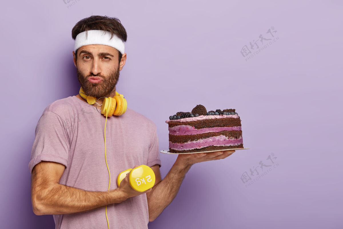 感觉积极认真的家伙有强壮的二头肌 拿着黄色的哑铃 过着健康的生活方式 拿着新鲜出炉的蛋糕姿势穿着盘子
