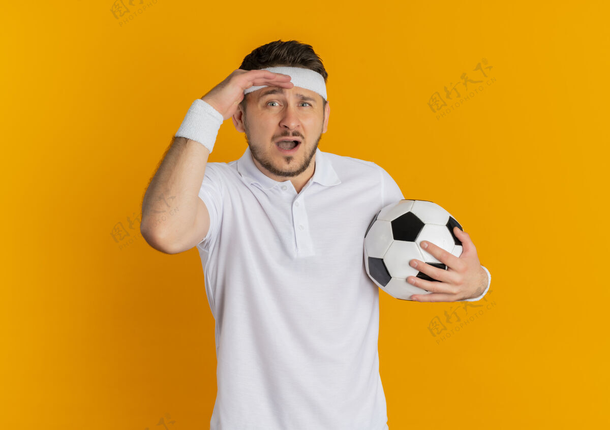 足球身穿白衬衫 头戴足球带的年轻健身男子站在橙色背景下困惑地看着摄像机橙色白色头带