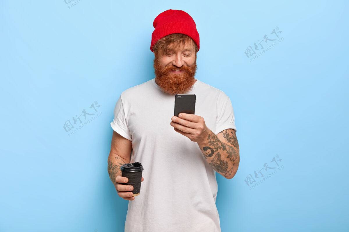 杯子积极向上的情感和现代科技理念开朗时尚的男人戴着红帽子和白t恤 留着姜黄色胡须 通过手机与朋友聊天 连接无线网络 喝咖啡红发手机胡须