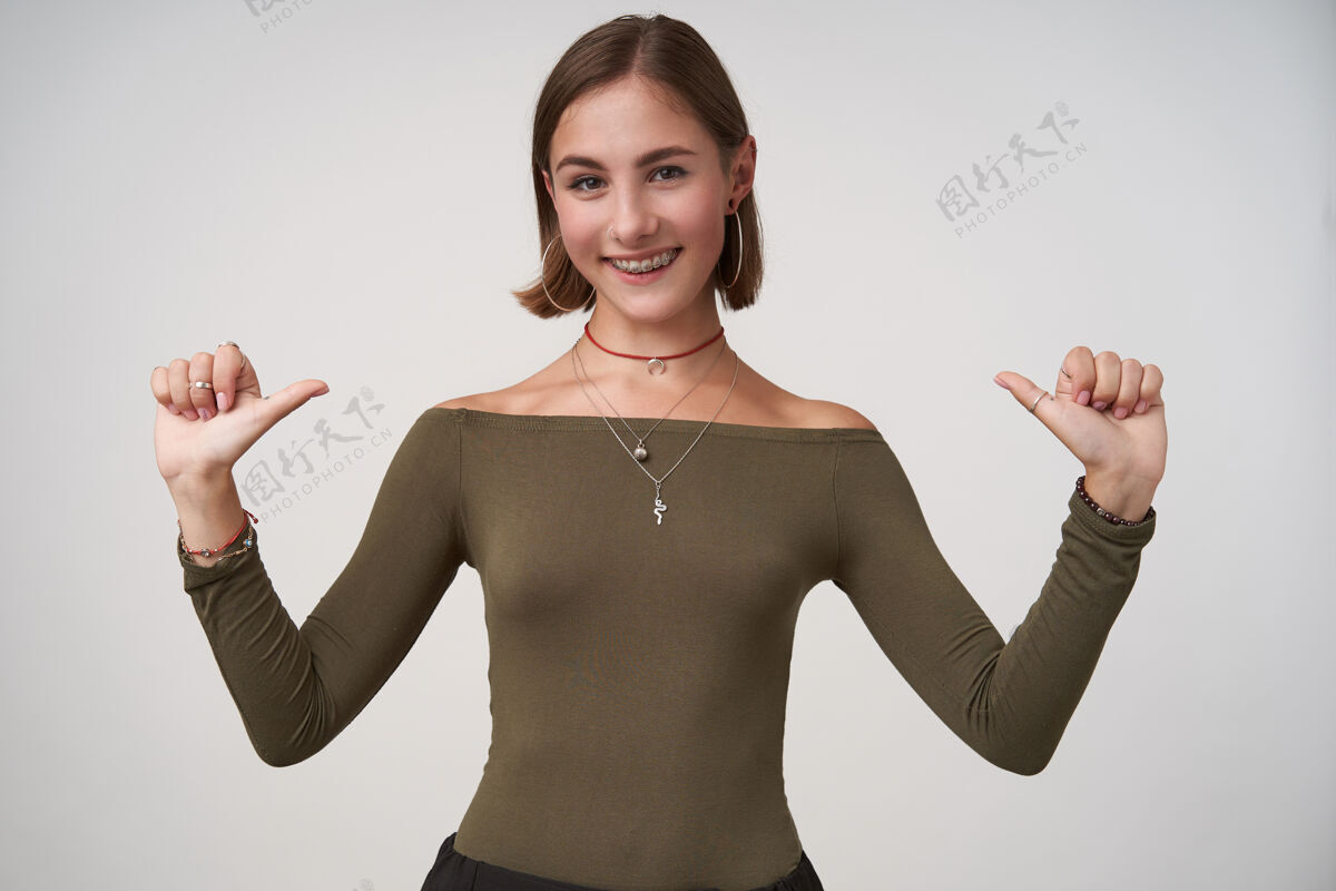 姿势快乐的年轻漂亮的黑发女子 随意的发型 穿着橄榄色上衣站在白墙上 高兴地用手指着自己发型时髦站