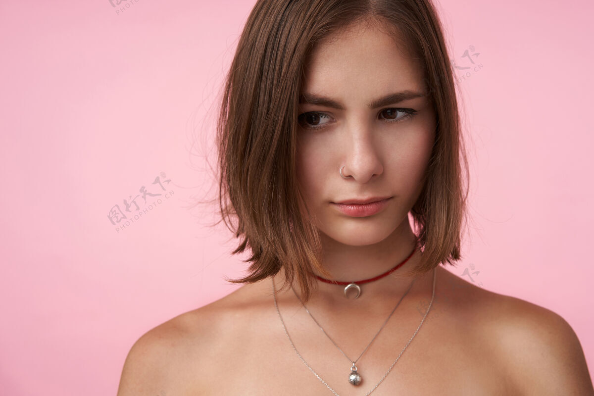 自然年轻而迷人的短发黑发女士 自然妆容 戴着时髦的饰物 赤裸着双肩在粉红色的墙上摆姿势年轻人特写配饰