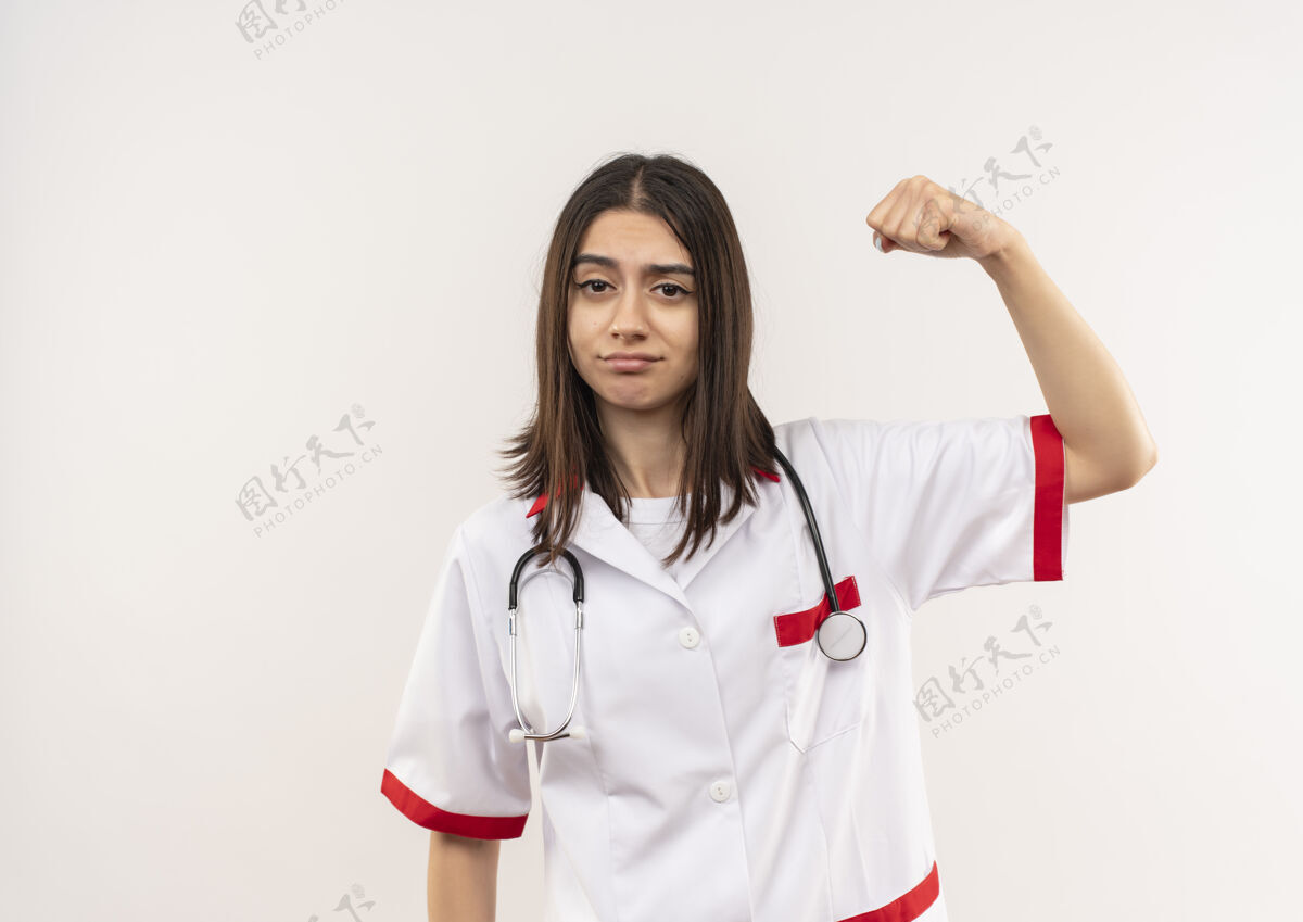 手势身穿白大褂的年轻女医生脖子上戴着听诊器握紧拳头举起手 看起来很自信 站在白墙上姿势握紧拳头
