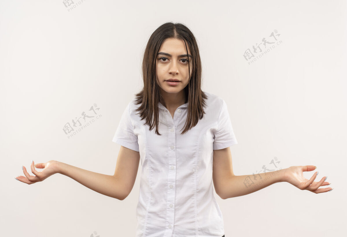 手臂穿着白衬衫的年轻女孩站在白墙上 神情茫然 两臂叉开 没有回答衬衫侧线姿势