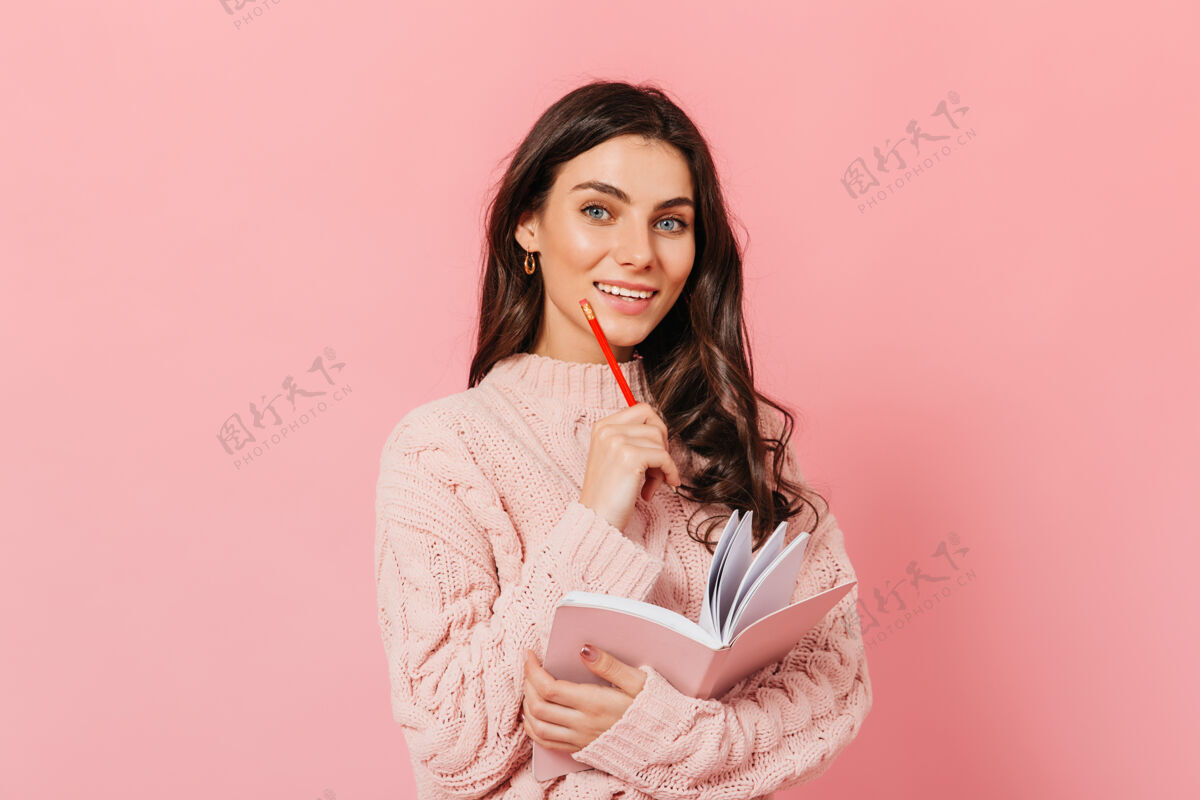 深色头发卷发女士带着雪白的微笑看着镜头穿着针织毛衣 手里拿着笔记本的女孩在孤立的背景上摆姿势女性日记粉色背景
