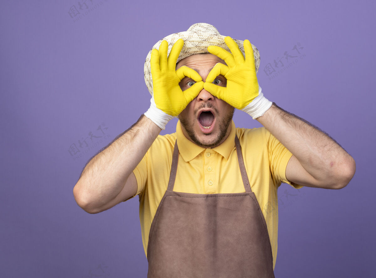 手指年轻的园丁穿着连身衣 戴着帽子 戴着工作手套 做着双目手势 手指透过手指看 站在紫色的墙上显得很惊讶男性工人工作