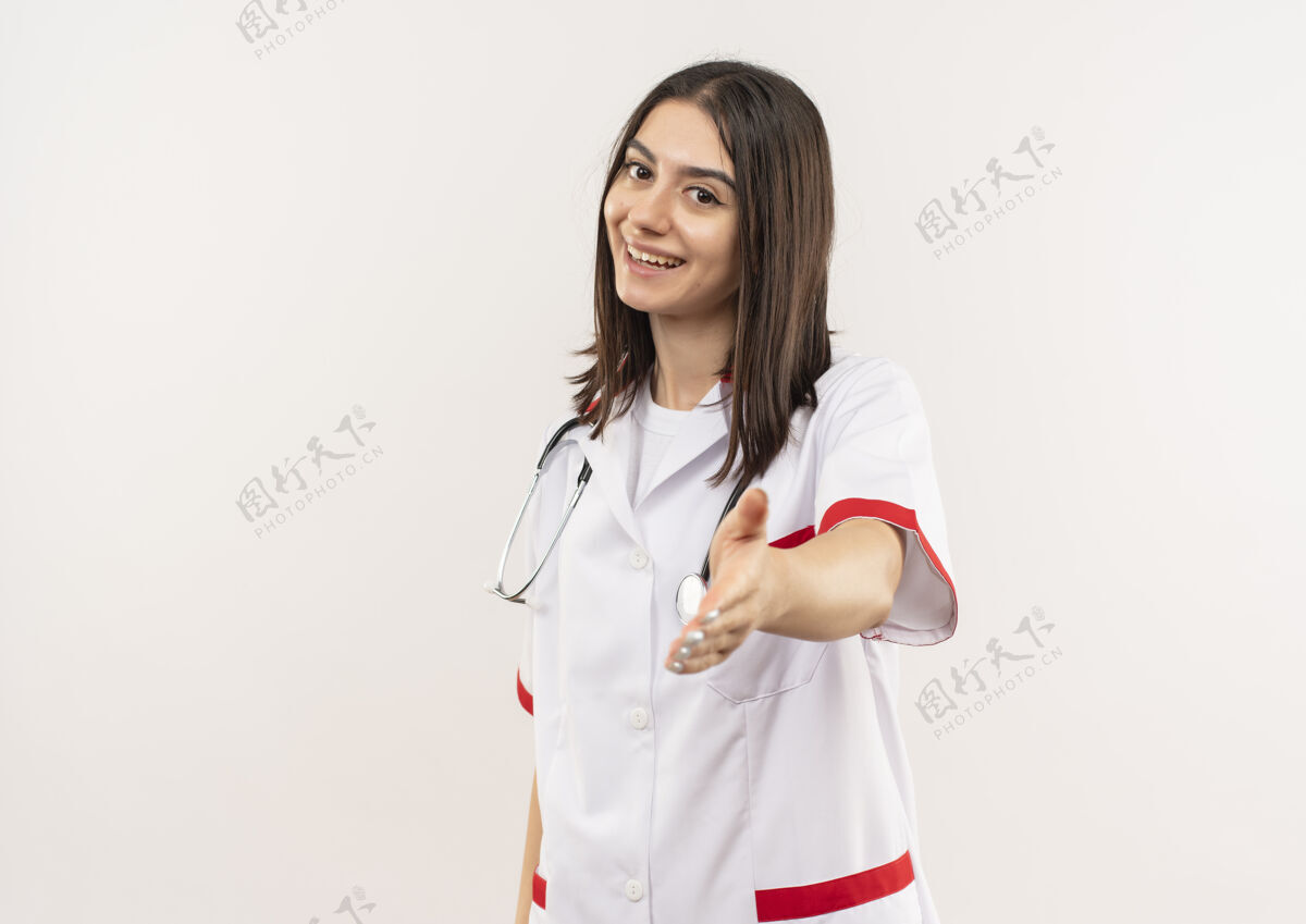看年轻的女医生穿着白大褂 脖子上戴着听诊器 面带微笑地看着前面 友好地伸出手 站在白墙上公民手势医生