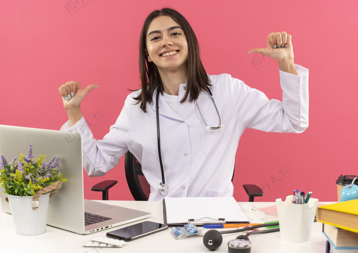 人年轻的女医生穿着白大褂 脖子上戴着听诊器 用手指指着自己 坐在桌旁 笔记本电脑盖在粉红色的墙上 看上去很满足手指医疗保健年轻人