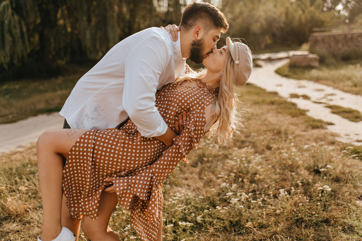 散步一个穿白衬衫的男人在温柔而热情的亲吻中倾斜着他的女朋友一对情侣在公园里拥抱拥抱棕色连衣裙女性